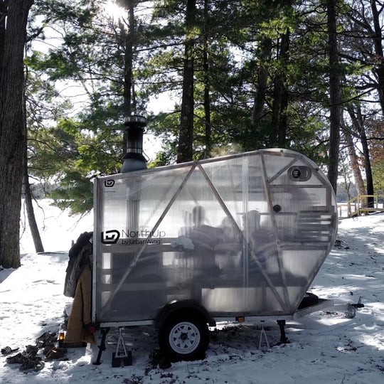 NorthUp Curve XL Mobile Sauna Trailer parked on a snowy lakeside landscape in front of pine trees with the sun peaking through.