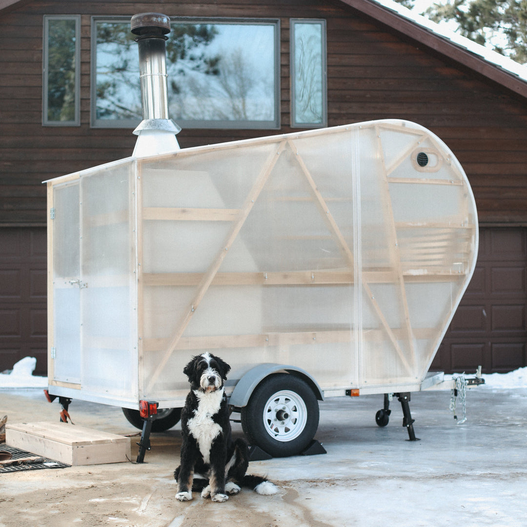 NorthUp XL mobile sauna trailer parked on an icy driveway in front of a cabin. Dog standing in front of the sauna trailer.